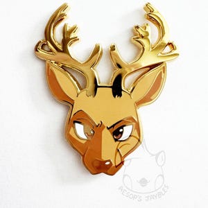 May include: A gold-toned metal enamel pin shaped like a deer head with large antlers. The deer features brown and tan enamel fill, dark eyes, and a stylized expression. A faint watermark logo with the text AESOP'S JAYBLES is visible below.