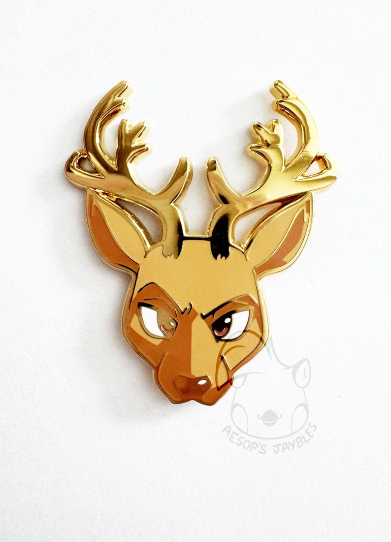 May include: A gold-toned metal enamel pin shaped like a deer head with large antlers. The deer features brown and tan enamel fill, dark eyes, and a stylized expression. A faint watermark logo with the text AESOP'S JAYBLES is visible below.
