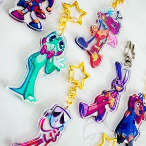 May include: A collection of acrylic keychains featuring colorful cartoon characters, including a green frog with a pink bow tie, a purple rabbit, and a jester. Each charm is attached to a gold-colored star-shaped clasp, with some designs displaying vibrant patterns and expressive faces.