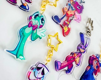 May include: A collection of acrylic keychains featuring colorful cartoon characters, including a green frog with a pink bow tie, a purple rabbit, and a jester. Each charm is attached to a gold-colored star-shaped clasp, with some designs displaying vibrant patterns and expressive faces.