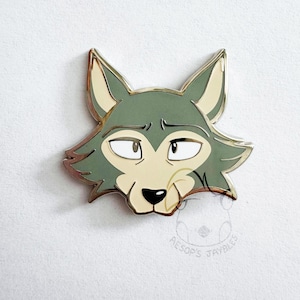 May include: A metal enamel pin featuring the stylized head of a wolf with sage green fur, cream-colored accents, and a black nose. The wolf has expressive eyes and pointed ears. A faint circular logo with the text AESOP'S JAYBLES is visible below.