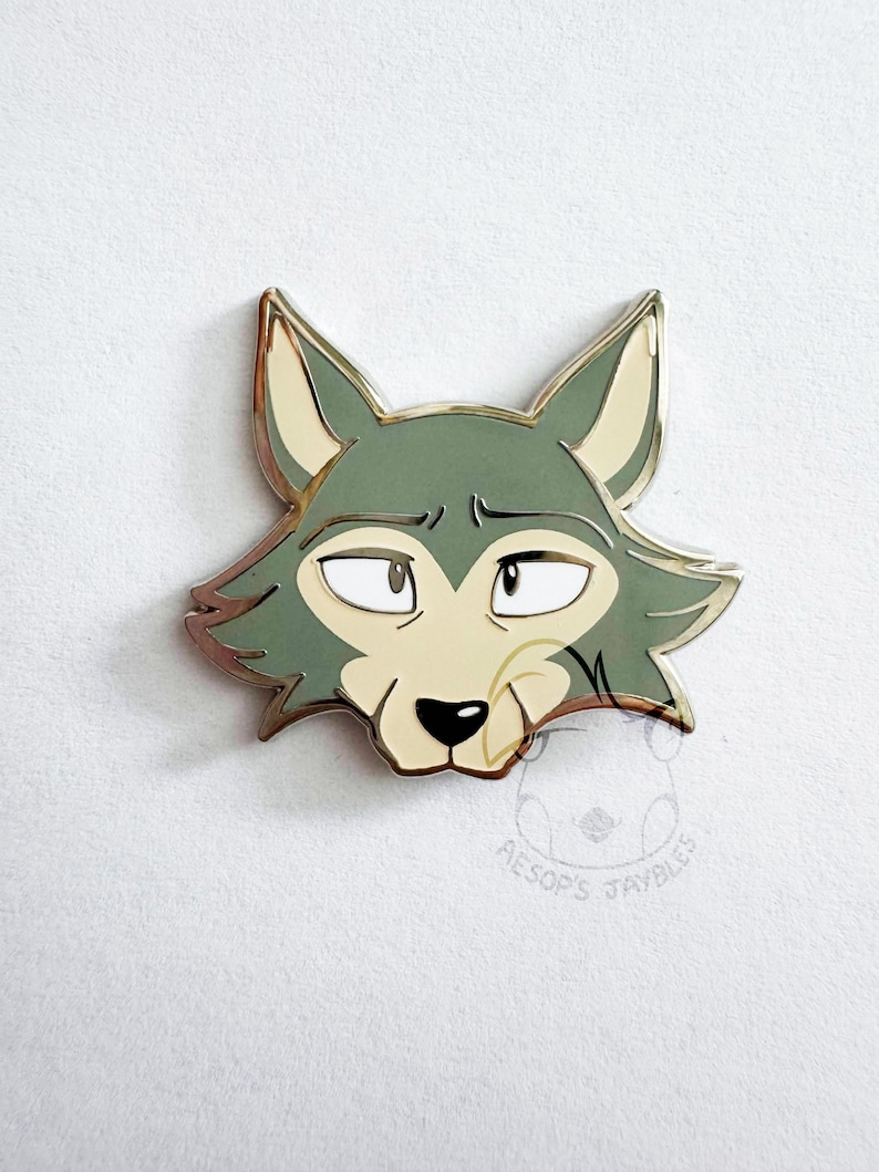 May include: A metal enamel pin featuring the stylized head of a wolf with sage green fur, cream-colored accents, and a black nose. The wolf has expressive eyes and pointed ears. A faint circular logo with the text AESOP'S JAYBLES is visible below.