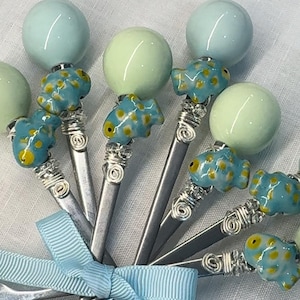 May include: Set of six silver teaspoons with blue and green glass beads and blue and yellow fish charms. The spoons are tied together with a light blue ribbon.