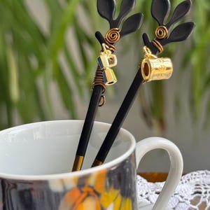Könnte beinhalten: Zwei dekorative Kaffeelöffel mit schwarzen Stielen und blattförmigen Oberteilen. Ein Löffel hat einen Miniatur-Gold-Kaffeekannen-Anhänger, der andere einen Gold-Kaffeemaschinen-Anhänger. Die Löffel befinden sich in einer weißen Tasse mit einem bunten Blumendesign.