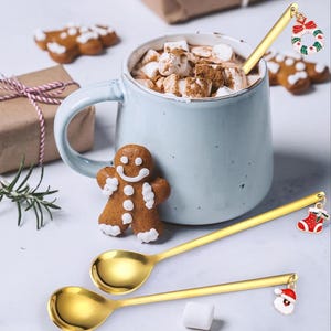 Könnte beinhalten: Eine hellblaue Tasse mit heißer Schokolade und Marshmallows, begleitet von zwei goldenen Löffeln mit Weihnachtsanhängern. Ein Lebkuchenmann und ein Lebkuchenkeks sind ebenfalls vorhanden, zusammen mit einem verpackten Geschenk.