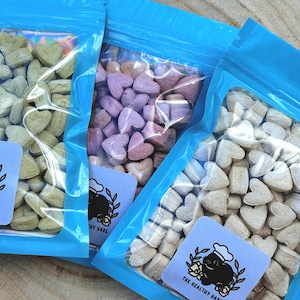 May include: Three clear plastic bags filled with heart-shaped treats in shades of green, purple, and beige. Each bag is sealed with a bright blue top. A small, light blue label with a cartoon hare wearing a chef's hat is visible on each bag.