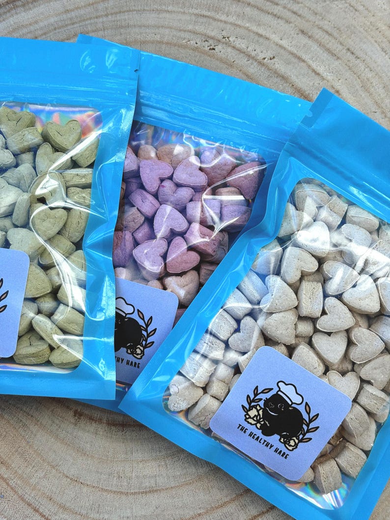 May include: Three clear plastic bags filled with heart-shaped treats in shades of green, purple, and beige. Each bag is sealed with a bright blue top. A small, light blue label with a cartoon hare wearing a chef's hat is visible on each bag.