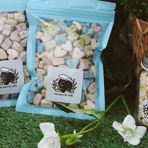 May include: Two clear plastic bags filled with heart-shaped, pastel-colored treats. Each bag has a light blue zipper. A jar of oats and a wicker basket are in the background. The bags have a sticker that says "THE HEALTHY HARE".