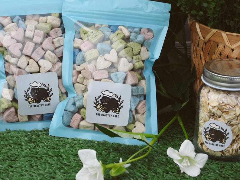 May include: Two clear plastic bags filled with heart-shaped, pastel-colored treats. Each bag has a light blue zipper. A jar of oats and a wicker basket are in the background. The bags have a sticker that says "THE HEALTHY HARE".