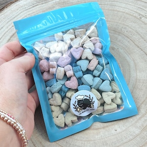 May include: A clear plastic bag with a light blue border filled with heart-shaped candies in pastel colors. A circular sticker with a chef pig graphic and the text "THE HEALTHY BAKE" is visible. The bag is held against a wooden background.