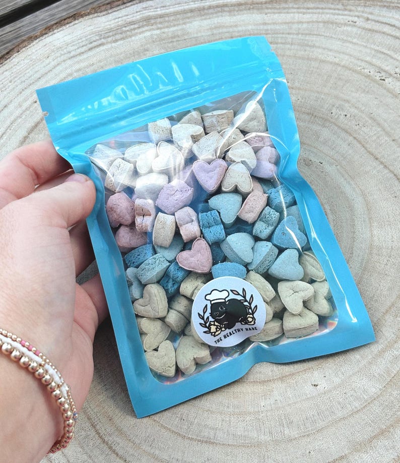 May include: A clear plastic bag with a light blue border filled with heart-shaped candies in pastel colors. A circular sticker with a chef pig graphic and the text "THE HEALTHY BAKE" is visible. The bag is held against a wooden background.