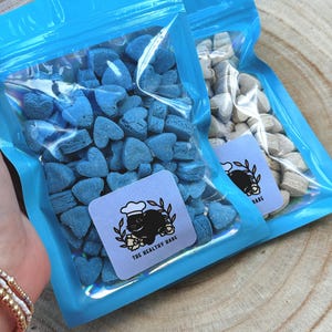 May include: Two clear plastic bags filled with heart-shaped treats. One bag contains blue treats, the other contains beige treats. Each bag has a blue label with a black cat wearing a chef's hat and the text "THE HEALTHY HARE".