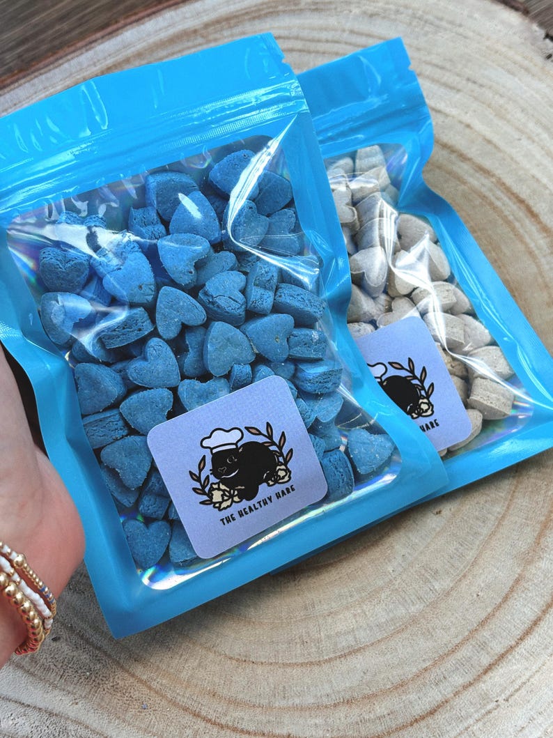 May include: Two clear plastic bags filled with heart-shaped treats. One bag contains blue treats, the other contains beige treats. Each bag has a blue label with a black cat wearing a chef's hat and the text "THE HEALTHY HARE".