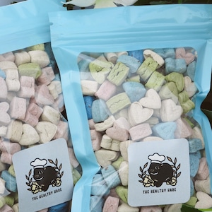 May include: Two clear plastic bags filled with colorful, heart-shaped and rectangular marshmallows. The marshmallows are pastel shades of pink, blue, green, and beige. Each bag has a light blue top and a square label with a cartoon rabbit wearing a chef's hat.