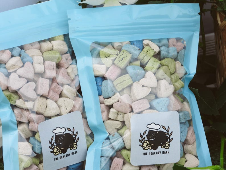 May include: Two clear plastic bags filled with colorful, heart-shaped and rectangular marshmallows. The marshmallows are pastel shades of pink, blue, green, and beige. Each bag has a light blue top and a square label with a cartoon rabbit wearing a chef's hat.