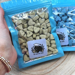 May include: Two clear plastic bags with blue zippers filled with heart-shaped treats. One bag contains light green treats, and the other contains blue treats. Each bag has a white label with a black illustration of a rabbit wearing a chef's hat and the text "THE HEALTHY HARE".