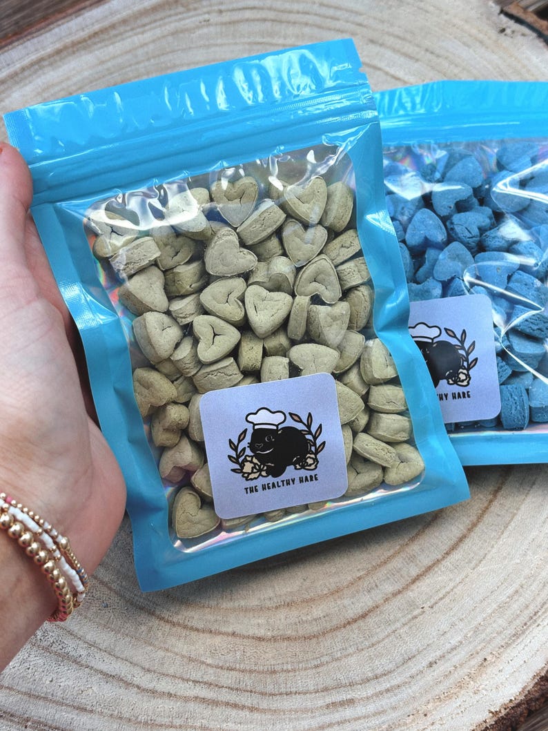 May include: Two clear plastic bags with blue zippers filled with heart-shaped treats. One bag contains light green treats, and the other contains blue treats. Each bag has a white label with a black illustration of a rabbit wearing a chef's hat and the text "THE HEALTHY HARE".