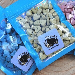 May include: Several clear plastic bags filled with heart-shaped treats in various colors. Each bag has a blue border and a square label with a black rabbit wearing a chef's hat and the text "THE HEALTHY HARE".