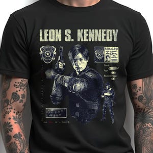May include: Black t-shirt featuring a graphic of Leon S. Kennedy holding a gun, with text that reads "LEON S. KENNEDY." The design includes police badges and a character ID card. The shirt is a crew neck.