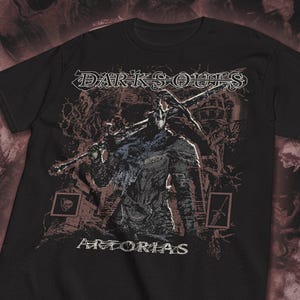 Könnte beinhalten: Ein schwarzes Kurzarm-T-Shirt mit einem Grafikdruck eines Ritters in dunkler Rüstung, der ein großes Schwert über der Schulter trägt. Der Text "DARK SOULS" steht oben und "ARTORIAS" unten in einer stilisierten weißen Schriftart.