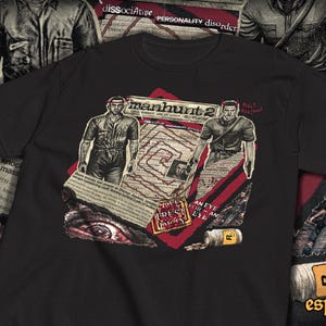May include: Black t-shirt featuring a graphic design with the text "manhunt 2". The design includes illustrations of two figures, newspaper clippings, and the text "LOVE TRUST PLAY". A Rockstar Games logo is visible.