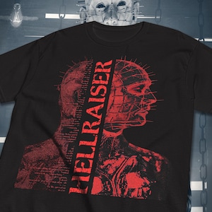 Peut inclure: T-shirt noir avec un motif rouge représentant une figure avec un motif quadrillé et le mot "HELLRAISER" en police verticale. Le design a un thème d'horreur, avec une illustration détaillée du profil d'un personnage.