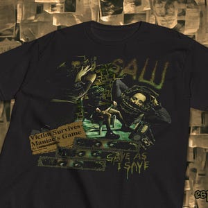 Peut inclure: T-shirt noir avec un motif graphique du film "Saw". Le motif comprend le mot "SAW" en vert, des images de personnages et le texte "Victim Survives Maniac's Game" et "SAVE AS I SAYE."