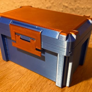 Könnte beinhalten: Eine blaue und orangefarbene rechteckige Box mit einem Klappdeckel und einem Verschluss. Die Box hat eine strukturierte Oberfläche und besteht aus Kunststoff. Der Deckel und der Verschluss sind orange, während der Körper der Box blau ist.