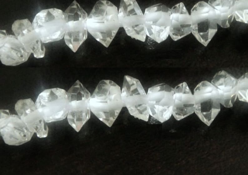 Op de afbeelding: Twee strengen heldere, facetgeslepen kristallen kralen. De kralen hebben een diamantachtige vorm en zijn geregen op een witte draad. De kristallen zijn transparant en reflecteren licht. De achtergrond is donker.