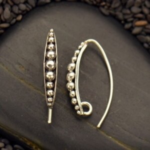May include: A pair of silver earrings. One earring is a vertical, elongated design with a series of silver beads. The other earring has a curved hook with a cluster of silver beads. The earrings are displayed on a dark, textured surface.