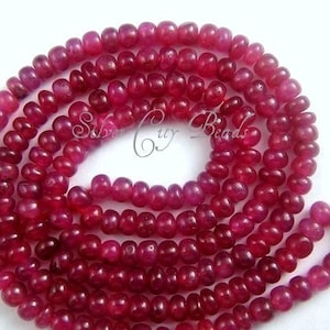 May include: A strand of deep red ruby beads, each bead is round and slightly elongated, creating a textured look. The beads are strung together to form a long necklace.