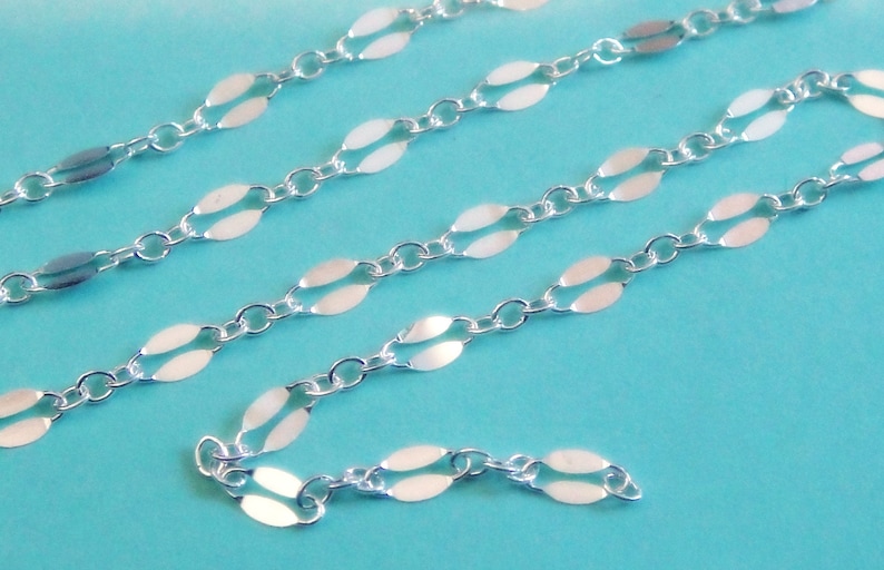 May include: Close-up of a delicate silver chain with oval-shaped links. The chain is displayed against a light blue background, showcasing its intricate design and reflective surface. Ideal for jewelry making or as a standalone accessory.