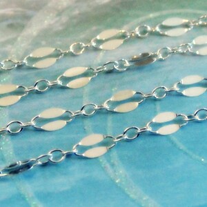 May include: Close-up of three strands of delicate silver chain. Each chain features a repeating pattern of small, oval-shaped links connected by tiny circular links. The chains are displayed against a soft, blue and white background.