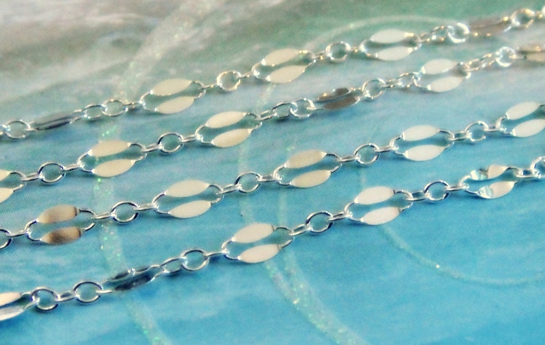 May include: Close-up of three strands of delicate silver chain. Each chain features a repeating pattern of small, oval-shaped links connected by tiny circular links. The chains are displayed against a soft, blue and white background.