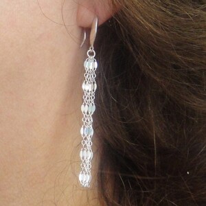 May include: A silver drop earring featuring a chain of small, flat, oval links. The earring is attached to a curved hook. The earring is shown against a background of brown hair and skin.