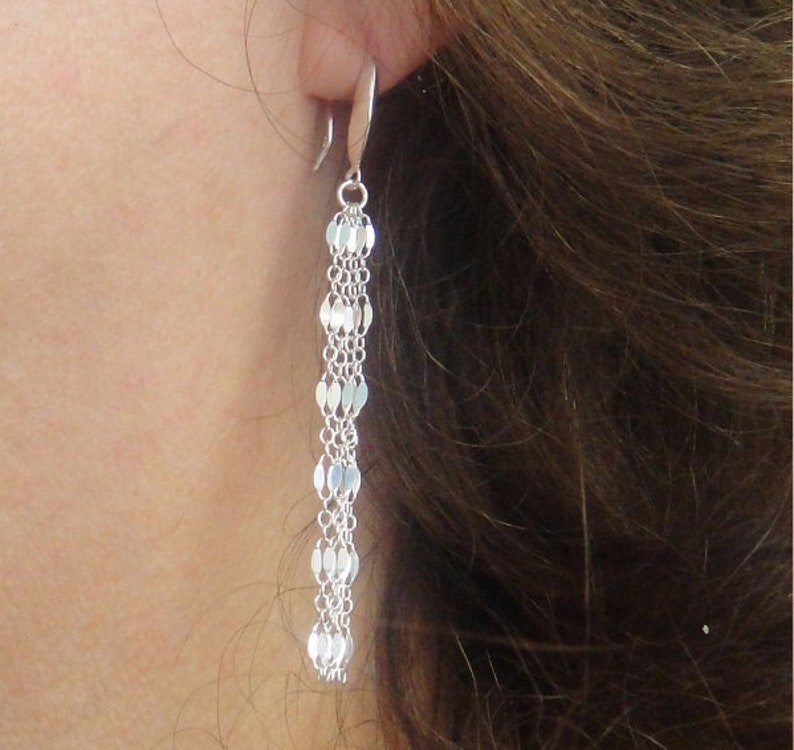 May include: A silver drop earring featuring a chain of small, flat, oval links. The earring is attached to a curved hook. The earring is shown against a background of brown hair and skin.