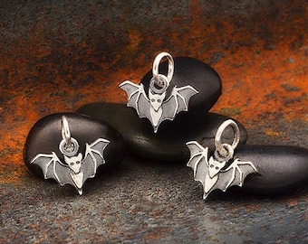 May include: Three silver bat charms with black wings and white faces. The charms are displayed on gray stones.