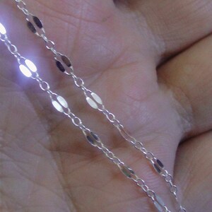 May include: Close-up of a delicate silver chain with oval-shaped links. The chain is thin and appears to be made of a shiny metal, reflecting light. The chain is likely a necklace or bracelet.