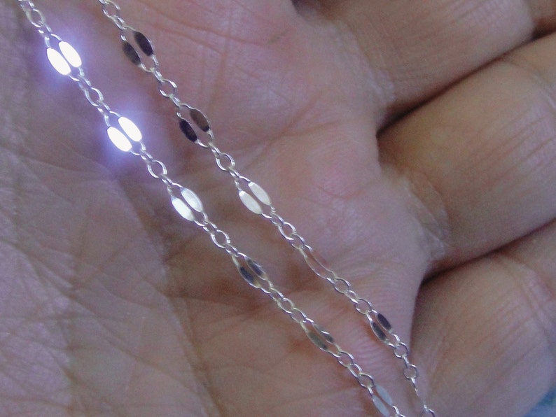 May include: Close-up of a delicate silver chain with oval-shaped links. The chain is thin and appears to be made of a shiny metal, reflecting light. The chain is likely a necklace or bracelet.