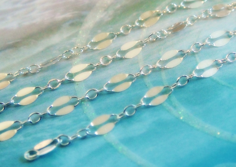 May include: Close-up of several strands of delicate silver chain. Each chain features oval-shaped links connected by small circular links. The chains are displayed against a soft, light blue and white background.