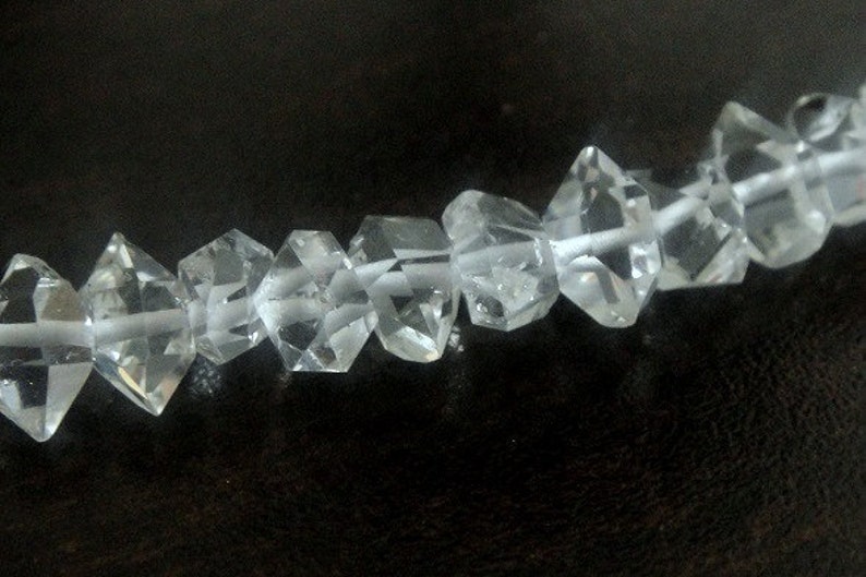 Op de afbeelding: Een streng heldere, facetgeslepen kristallen kralen. Elke kraal is diamantvormig met meerdere facetten, geregen op een dun, wit koord. De kralen zijn transparant, reflecteren licht en staan tegen een donkere achtergrond.