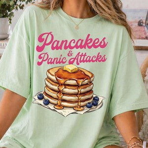 Può includere: T-shirt verde chiaro con la scritta rosa "Pancakes & Panic Attacks". La maglietta presenta una grafica di una pila di pancake con sciroppo, burro e mirtilli.