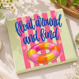 Könnte beinhalten: Ein hellgrünes, gefaltetes Handtuch mit einer Grafik eines rosa und gelben aufblasbaren Rings. Das Handtuch hat rosa vertikale Streifen und die Worte "Float around and find" in blauer Schrift.