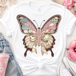 Peut inclure: Un t-shirt blanc à manches courtes avec un grand motif central représentant un papillon. Les ailes du papillon sont composées de divers motifs patchwork incluant des fleurs, du vichy et du tartan dans des tons rose, bleu et crème, avec une croix rose.