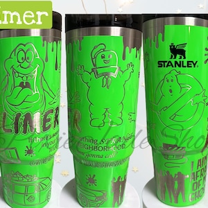 May include: Green stainless steel travel mug with a black lid and handle. The mug features a graphic of Slimer from Ghostbusters, with the text "Slimer" and "If there's something strange in your neighborhood, who ya gonna call?" printed on the side.