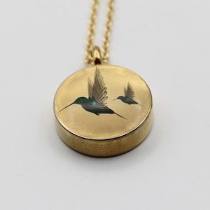 Puede incluir: Collar con colgante dorado con un dije redondo con dos colibríes en vuelo. Las aves están representadas con detalles verdes y negros. El collar tiene una cadena delicada y un cierre sencillo.