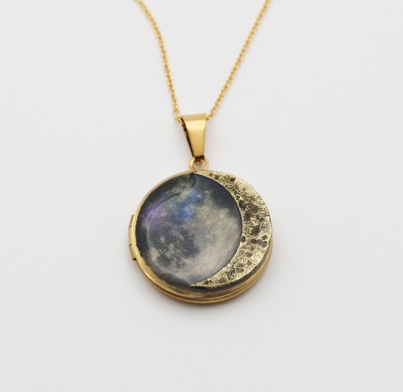 Puede incluir: Un collar de cadena dorada con un medall&oacute;n en forma de media luna. El medall&oacute;n tiene un dise&ntilde;o de galaxia con una media luna dorada en la parte delantera.