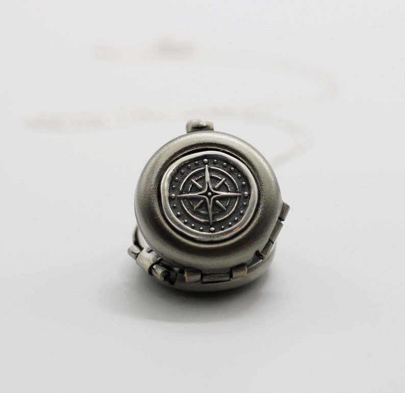 May include: A silver compass charm with a detailed design. The compass is attached to a silver chain.