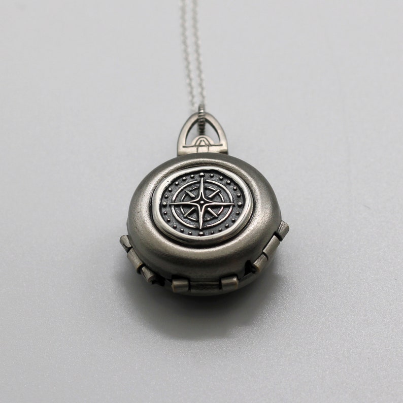 May include: A silver locket necklace with a compass design. The compass has a black face with a silver star in the center.