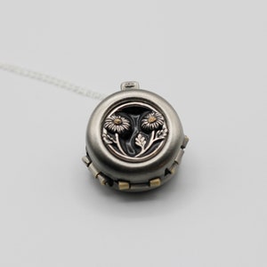 Peut inclure: Un collier pendentif rond argent&eacute; avec un motif floral. Le pendentif pr&eacute;sente deux fleurs d&eacute;taill&eacute;es sur un fond sombre. Le collier a une cha&icirc;ne d&eacute;licate et est pos&eacute; sur un fond blanc.